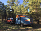 Camping in style! An orange SUV tows a blue pop-top Cricket camper in a sunny pine forest. Consider this RV rental for your next trip.
