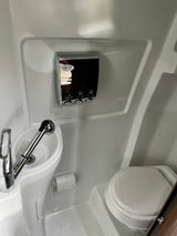 A compact white RV rental bathroom, featuring a toilet, small sink/shower, and wall-mounted dispensers.