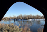 Scenic winter river view through an opening, from an RV rental. Bare trees, snow-dusted banks & calm water reflect the clear blue sky.