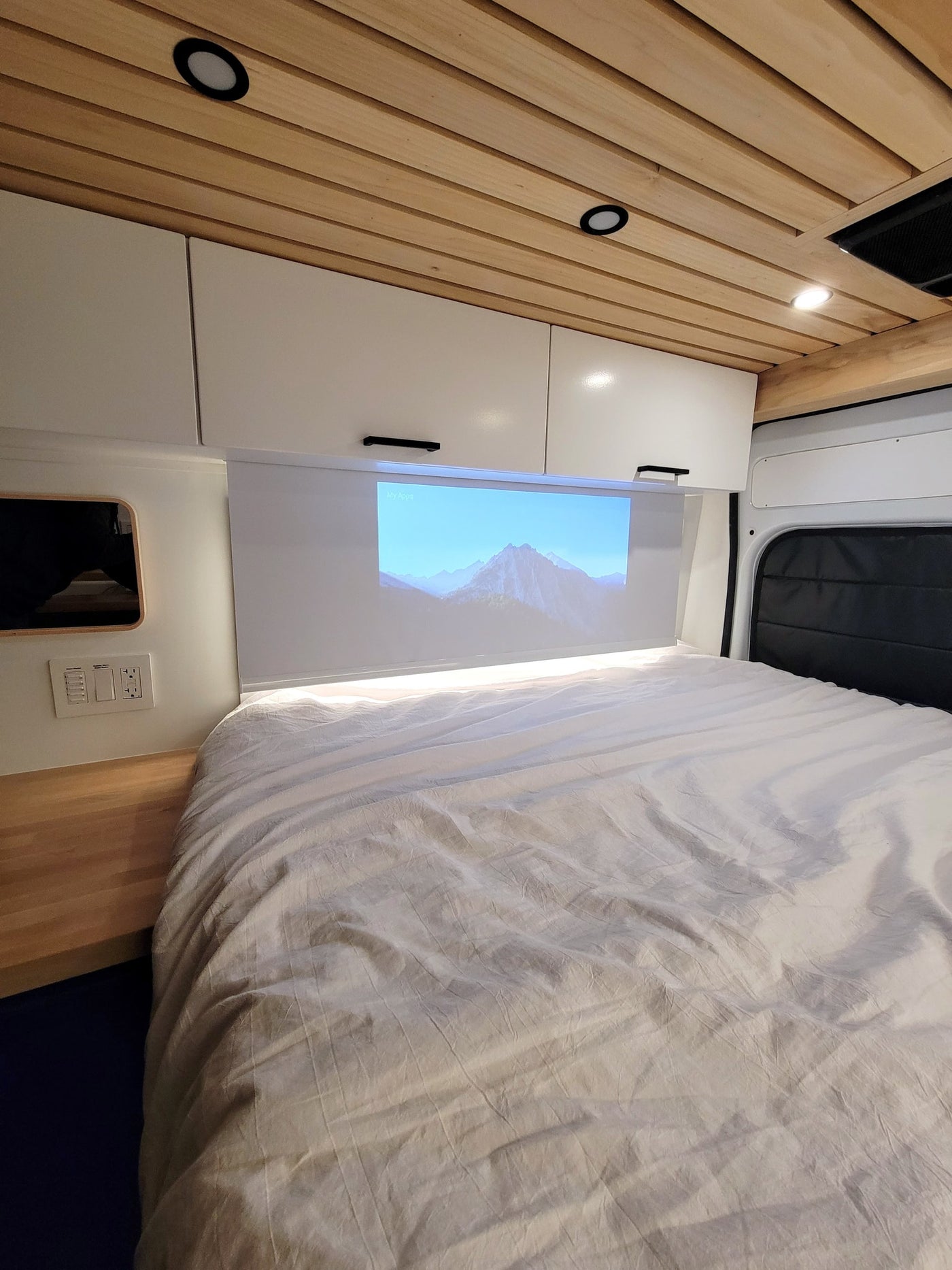 Cozy RV rental bedroom featuring a large bed, overhead cabinets, and a projector displaying mountains on the screen above. — photo 11