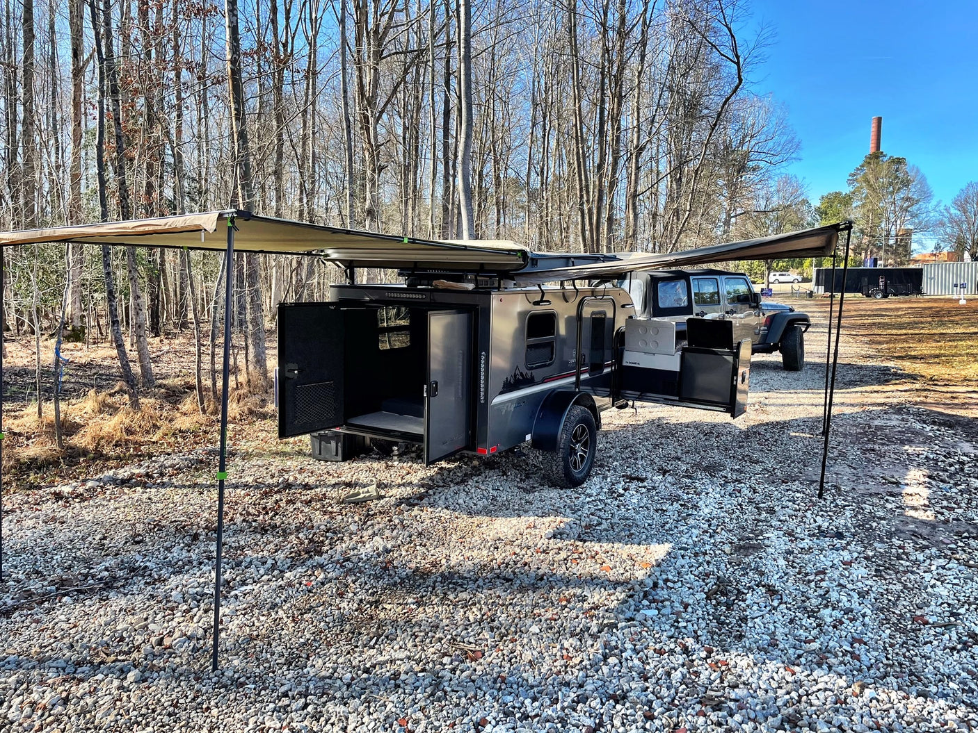 Compact RV rental: a dark camper trailer with dual awnings extended, parked on gravel with a Jeep, ready for outdoor adventure.