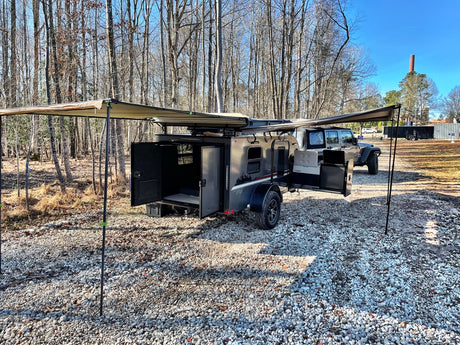 Compact RV rental: a dark camper trailer with dual awnings extended, parked on gravel with a Jeep, ready for outdoor adventure.