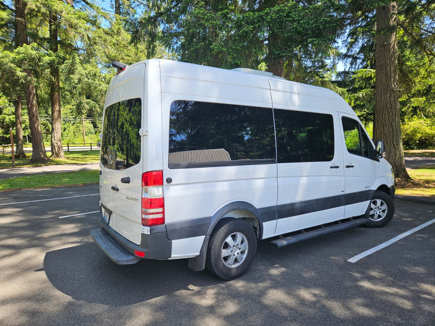 White Mercedes Sprinter van, an excellent RV rental option, parked under trees on a sunny day. Features tinted windows & a rear step.