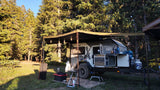 An RV rental, a compact teardrop camper, is set up with an outdoor kitchen in a sunny forest clearing.