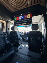Inside a luxurious rv rental: a large overhead TV displays live sports, surrounded by plush seating and a bright exterior view.