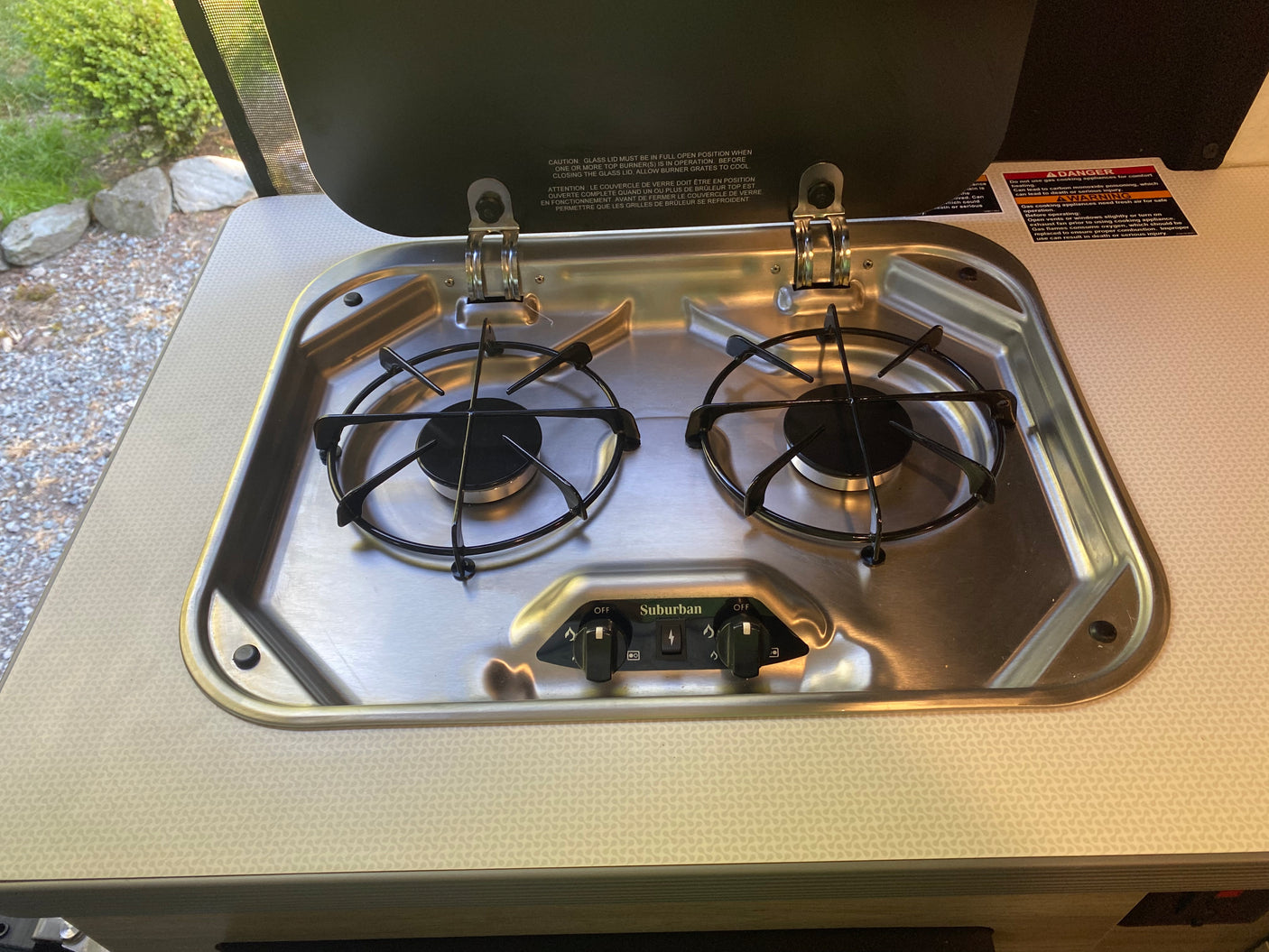 Close-up of a stainless steel Suburban gas cooktop in an RV rental, with two burners and safety warnings on the lid.