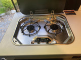 Close-up of a stainless steel Suburban gas cooktop in an RV rental, with two burners and safety warnings on the lid.