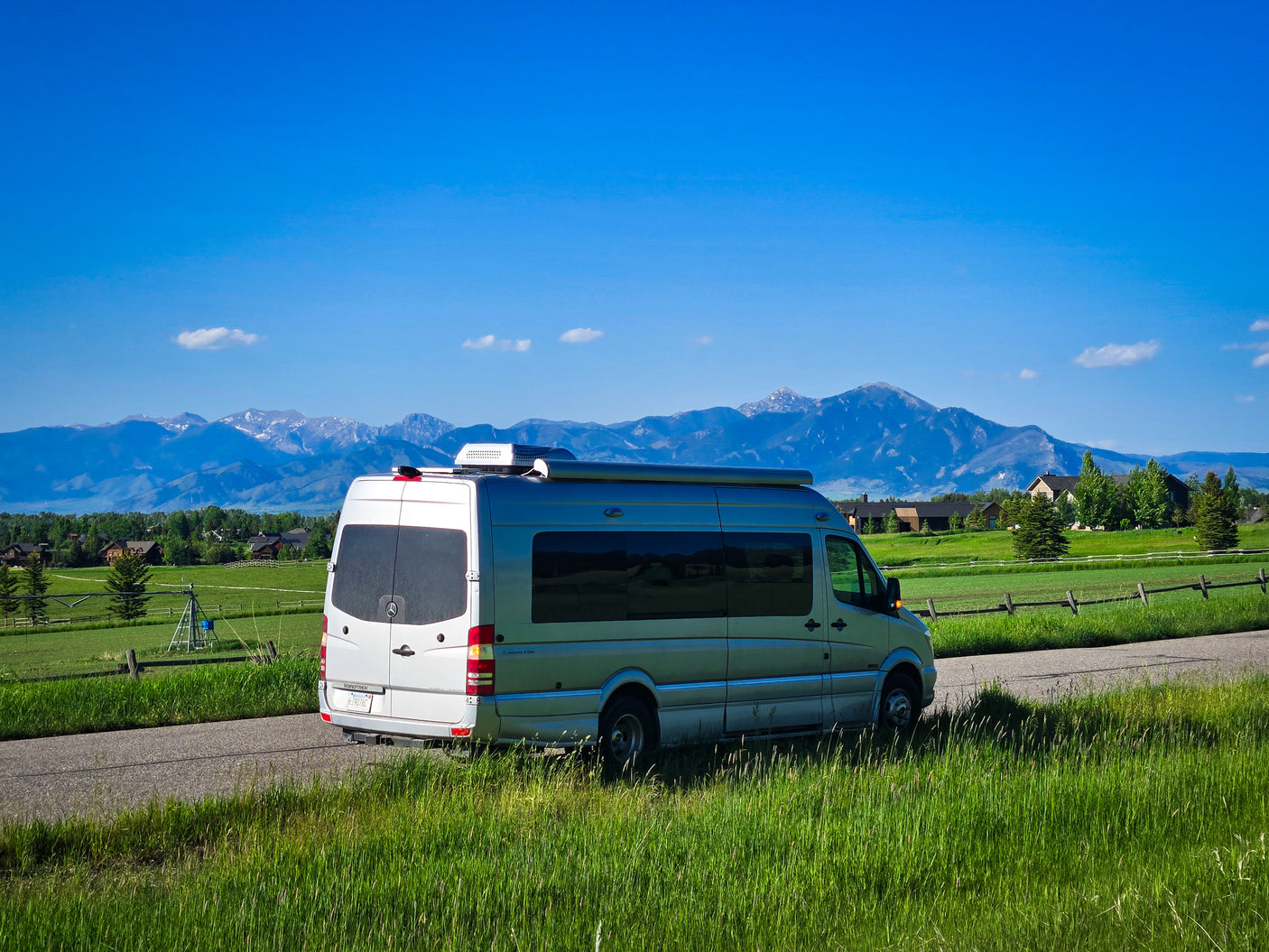 An RV rental (silver camper van) parked on a country road, framed by vibrant green fields and majestic snow-capped mountains.