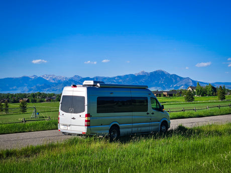 An RV rental (silver camper van) parked on a country road, framed by vibrant green fields and majestic snow-capped mountains.