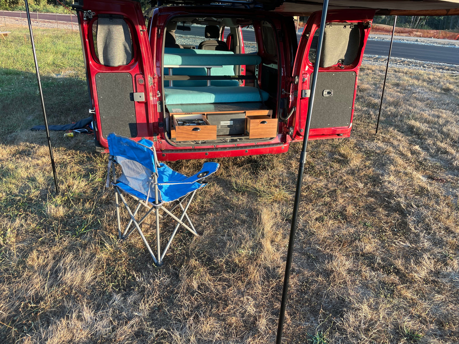 Red camper van with open rear doors reveals a cozy interior bed, pull-out kitchen & a camping chair. Great RV rental setup!