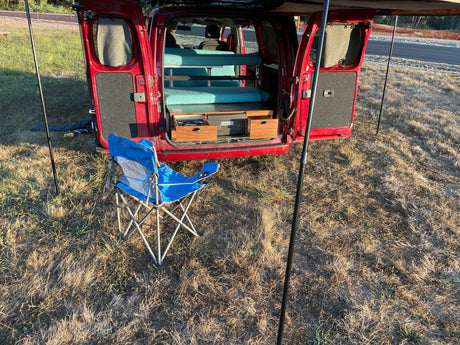 Red camper van with open rear doors reveals a cozy interior bed, pull-out kitchen & a camping chair. Great RV rental setup!