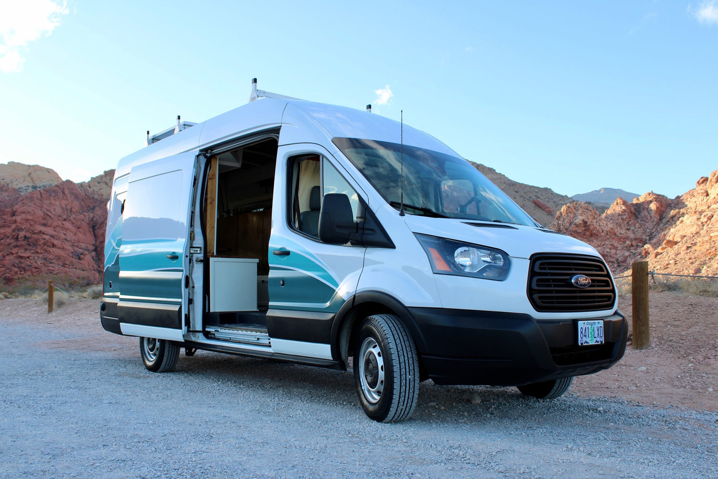 White Ford Transit rv rental camper van on gravel, side door open, amidst red rock mountains under a blue sky. — photo 11