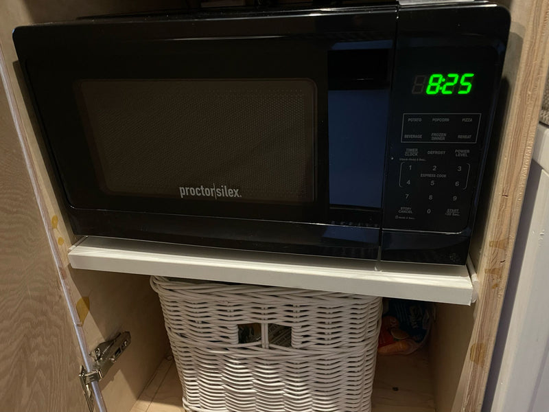 A black Proctor Silex microwave, displaying 8:25, built into an RV rental kitchen cabinet above a white wicker basket. — photo 16