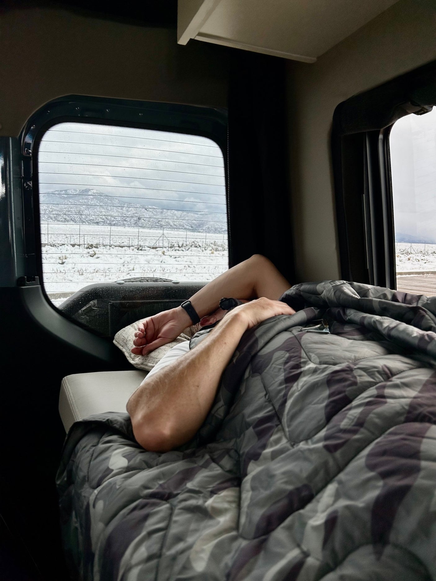Inside an RV rental: person covered by a blanket, peacefully napping with a snowy mountain landscape visible through the window.