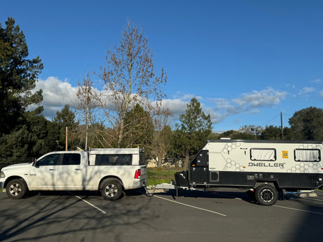 A white Ram pickup truck is towing a Dweller 15' travel trailer in a paved lot. A good look for an RV rental.