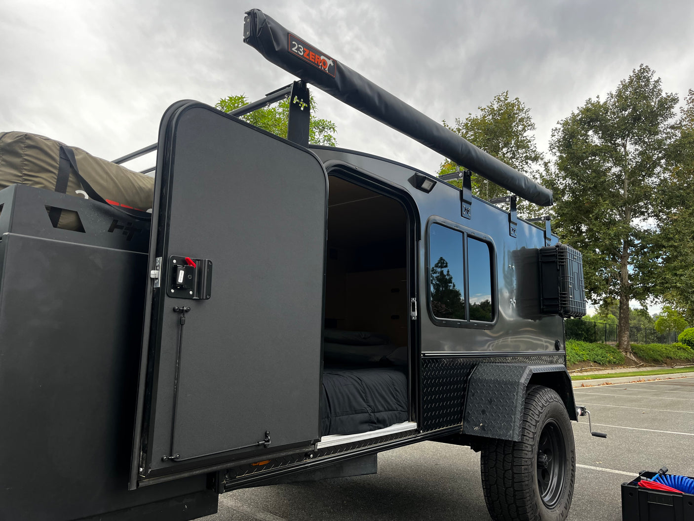 This dark gray off-road teardrop camper, shown with its door open revealing a cozy bed, is an ideal RV rental for outdoor adventures.