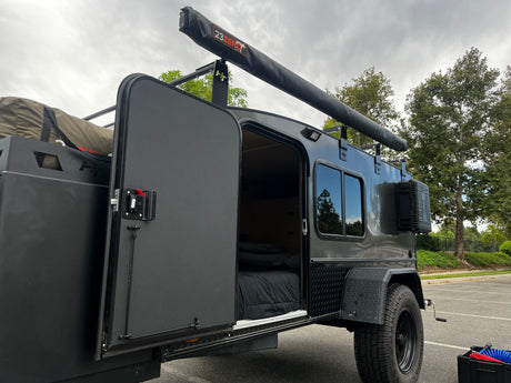 This dark gray off-road teardrop camper, shown with its door open revealing a cozy bed, is an ideal RV rental for outdoor adventures.