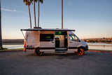 Experience comfort in this accessible rv rental camper van. Awning extended, door open, parked lakeside at golden hour.