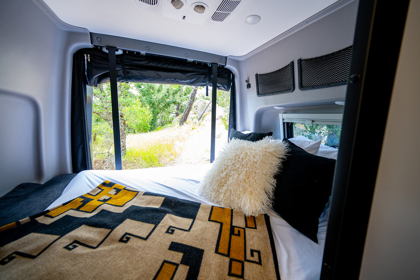 Bedroom of an rv rental showcasing a cozy bed with decorative blanket, scenic window view, and practical overhead storage nets.
