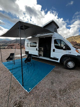 Scenic RV rental setup: white camper van with pop-top, extended awning, blue mat, table, & chairs amidst red rocks.