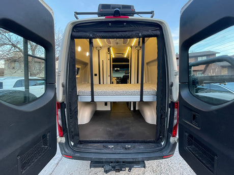 Rear interior of an RV rental camper van, showing an elevated bed, storage space, and bright LED lighting for van life.