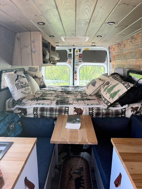 Cozy RV rental camper van interior: nature-themed bed, wooden ceiling/walls, kitchen, and dining nook for comfortable travel.