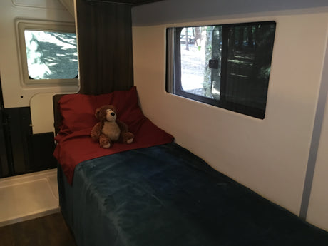 Cozy RV rental bedroom with a teddy bear on the bed and nature views from windows.
