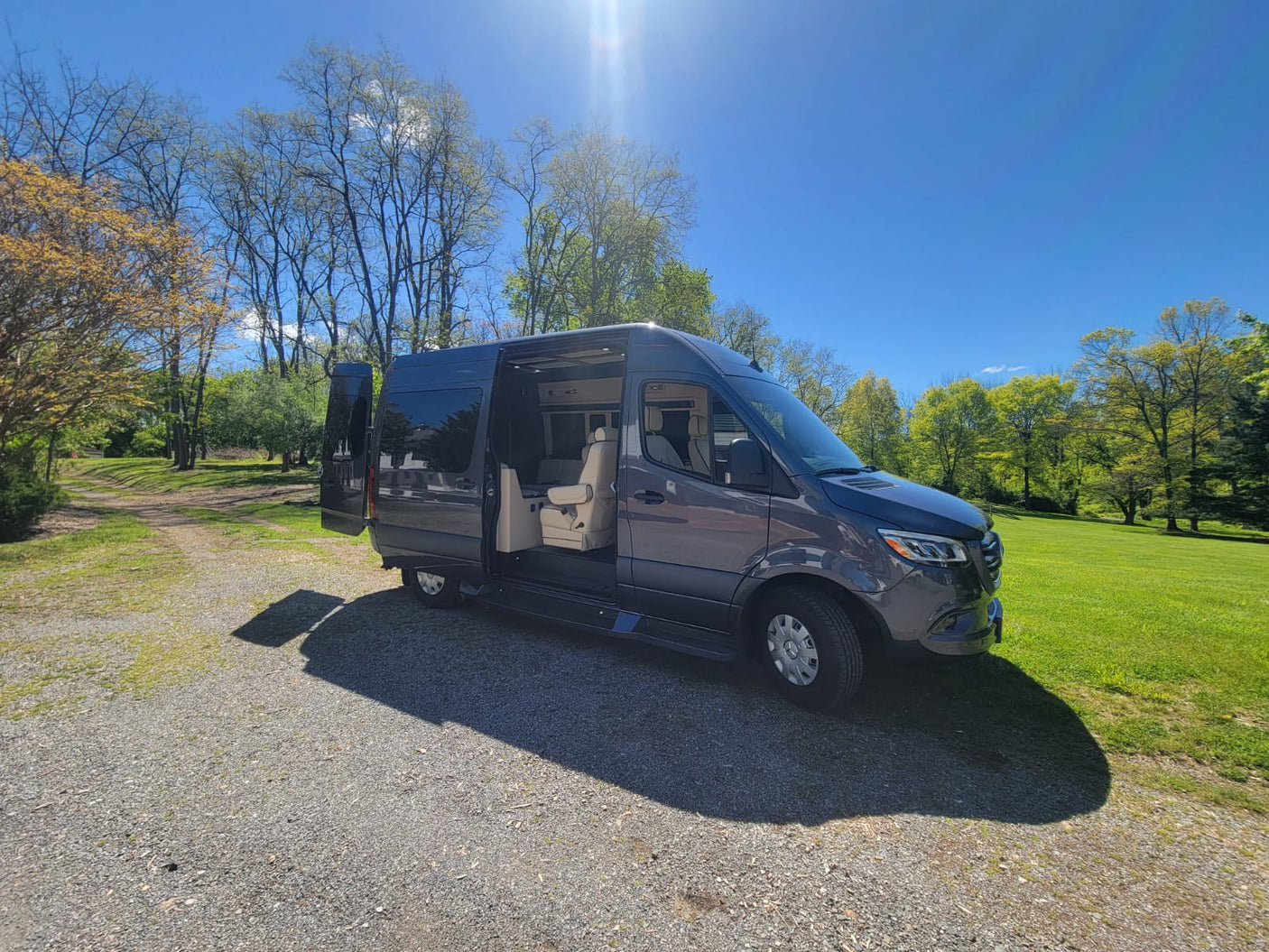 Gray RV rental camper van with open doors on gravel drive, revealing light interior. Parked by green trees and grass under a blue sky.