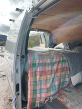 An RV rental camper van's interior: a bed with a colorful plaid blanket, wood ceiling, and open door view of mountains.