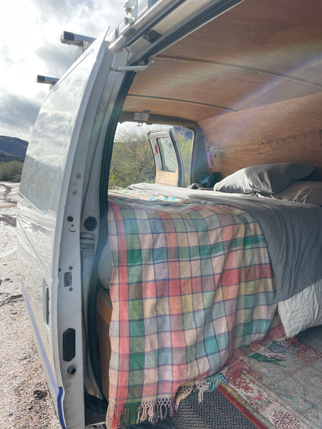 An RV rental camper van's interior: a bed with a colorful plaid blanket, wood ceiling, and open door view of mountains.