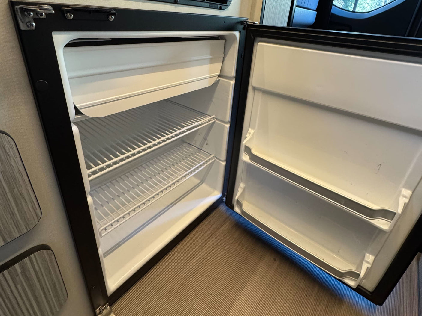 This clean, empty RV rental refrigerator, with its door open, shows ample wire shelves and door storage, ready for your trip's groceries.