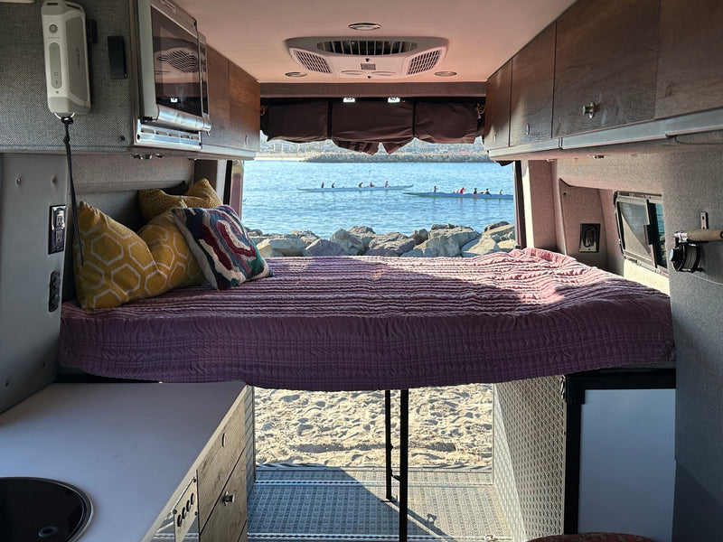 Inside an RV rental camper van: a cozy bed with purple quilt & pillows, overlooking the ocean, sandy beach, and canoes. — photo 13