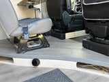 RV rental interior: Rear grey passenger seat, front black swivel seat, and a Lagun table mount. Light wood-look flooring with heater vent.