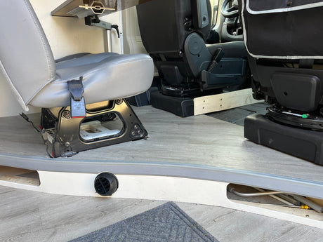 RV rental interior: Rear grey passenger seat, front black swivel seat, and a Lagun table mount. Light wood-look flooring with heater vent.
