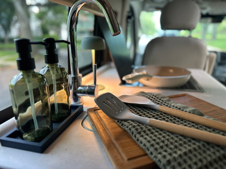 Green soap bottles, chrome faucet, spatulas on a cutting board in an RV rental kitchenette, ready for travel meals.