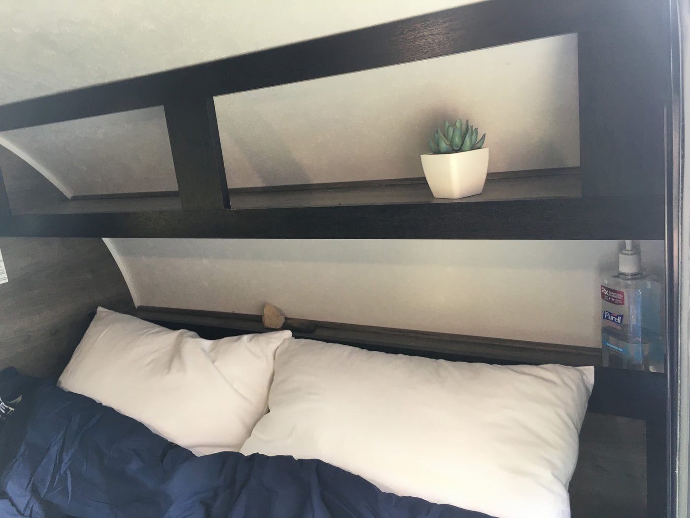 An RV rental's cozy bedroom features a bed with white pillows & blue blanket, plus overhead storage with a plant & Purell. — photo 6