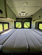 Inside a comfortable rv rental campervan, showing a large bed, overhead cabinets, and bright windows with views of green fields.