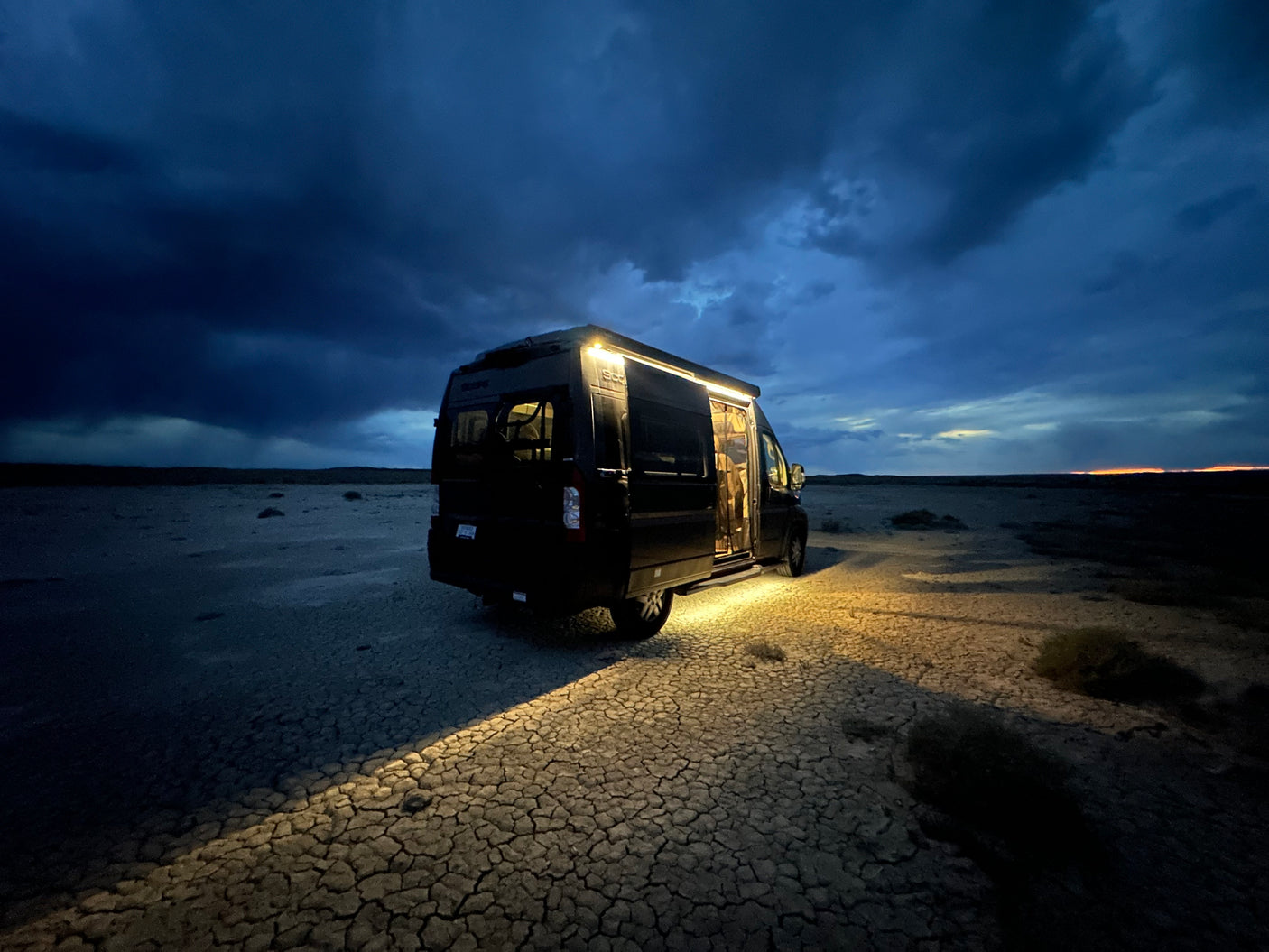 An RV rental van glows on cracked desert ground under dramatic dark blue twilight clouds. Ideal for off-grid adventure.