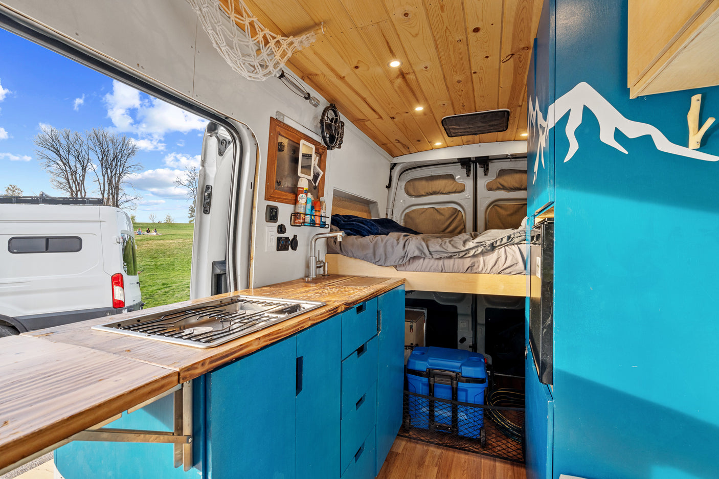 Stylish rv rental camper van interior: blue kitchen, wood ceiling, cozy bed, & park view perfect for van life adventures.