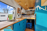Stylish rv rental camper van interior: blue kitchen, wood ceiling, cozy bed, & park view perfect for van life adventures.