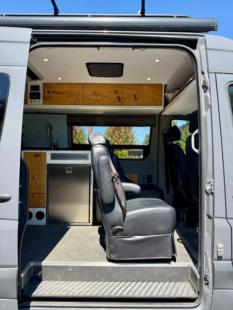 Explore this custom RV rental interior: modern camper van with built-in fridge, wood cabinetry, and swivel seating.