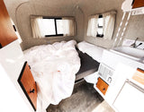 Compact rv rental interior with a cozy bed, white bedding, small sink, and windows providing natural light.