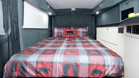 Cozy RV rental interior featuring a bed with plaid, bear-themed bedding, a white window blind, and storage cabinets.