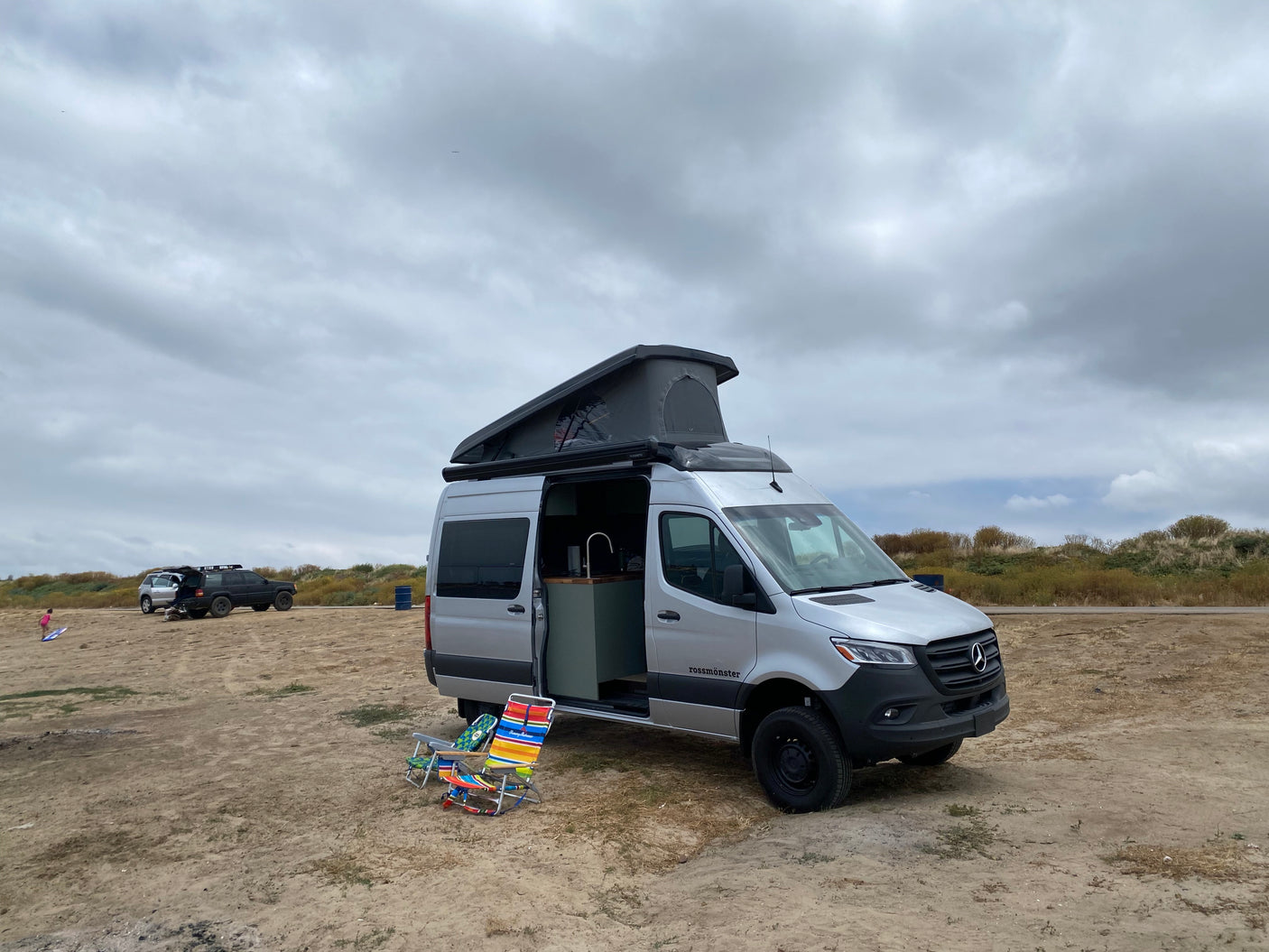 Silver Mercedes Sprinter RV rental camper van with pop-top, parked on a sandy area with chairs, other vehicles, under cloudy skies.