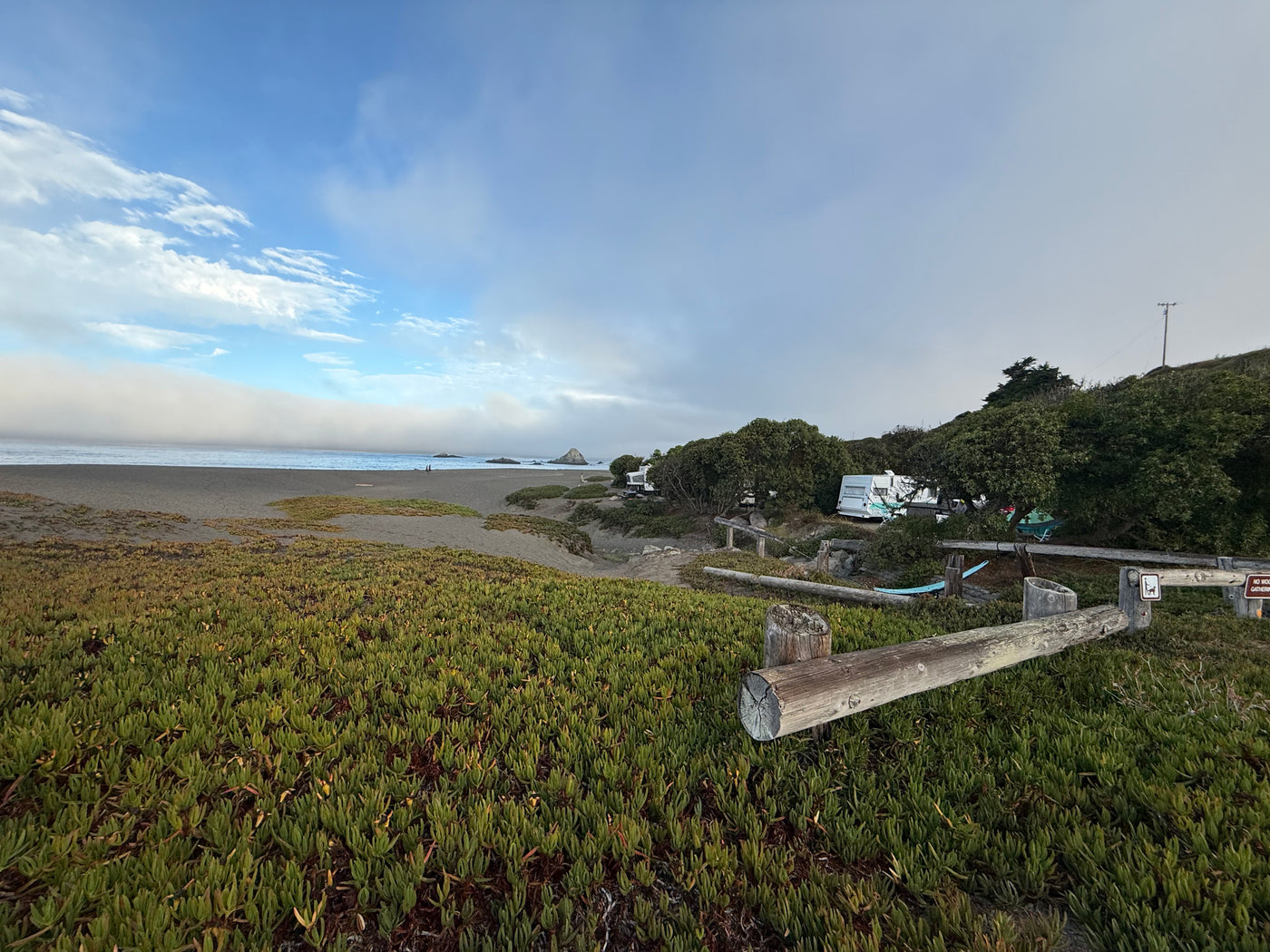RV rental trailers at a coastal campsite near a beach with ocean fog. Lush green foreground plants and wooden fence. — photo 18