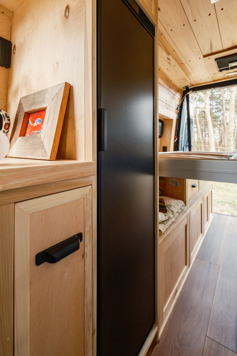Inside an rv rental, showcasing custom wood interior with a tall black refrigerator, cabinetry, and a view to nature. — photo 13