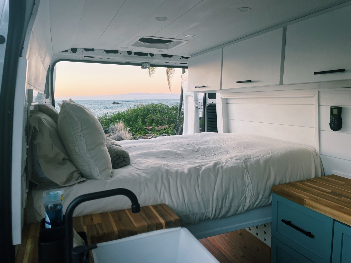 Cozy RV rental interior with a comfy bed, kitchenette, and white cabinets, offering stunning ocean views at sunset.