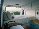 Cozy RV rental interior with a comfy bed, kitchenette, and white cabinets, offering stunning ocean views at sunset.