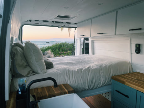 Cozy RV rental interior with a comfy bed, kitchenette, and white cabinets, offering stunning ocean views at sunset.
