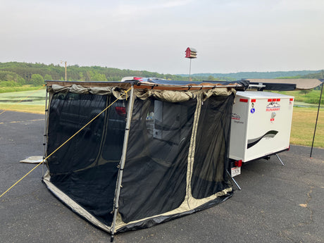 A compact Runaway RV rental, a white camper trailer, is set up with a large mesh screen room attached to a vehicle on asphalt. Ideal for camping.